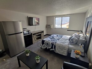 Habitación de hotel compacta con cama, escritorio pequeño, refrigerador, televisor y ventana con vista a la montaña.