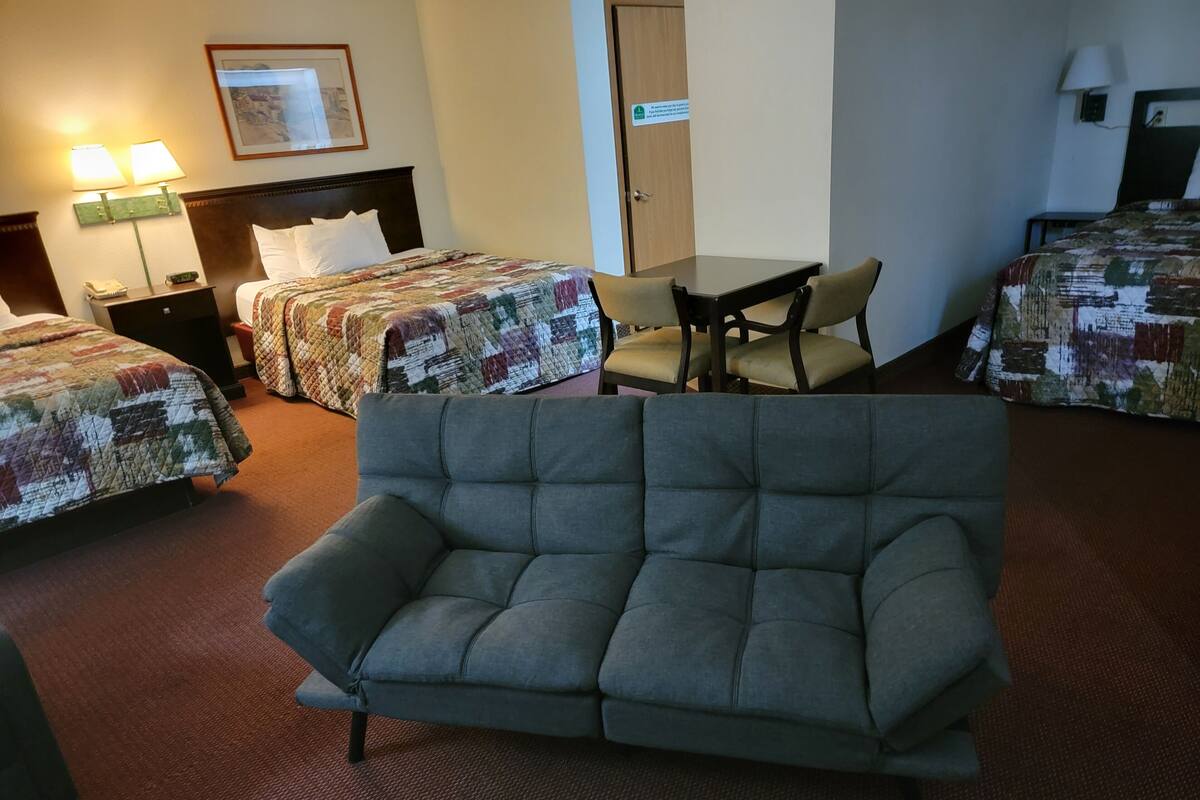A hotel room with a sofa, two beds with quilts, a table, chairs, a lamp, a phone, and a door.