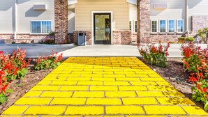 A building entrance with a yellow brick pathway leading to a door, surrounded by red flowers and a brick wall.