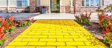 A building entrance with a yellow brick pathway leading to a door, surrounded by red flowers and a brick wall.