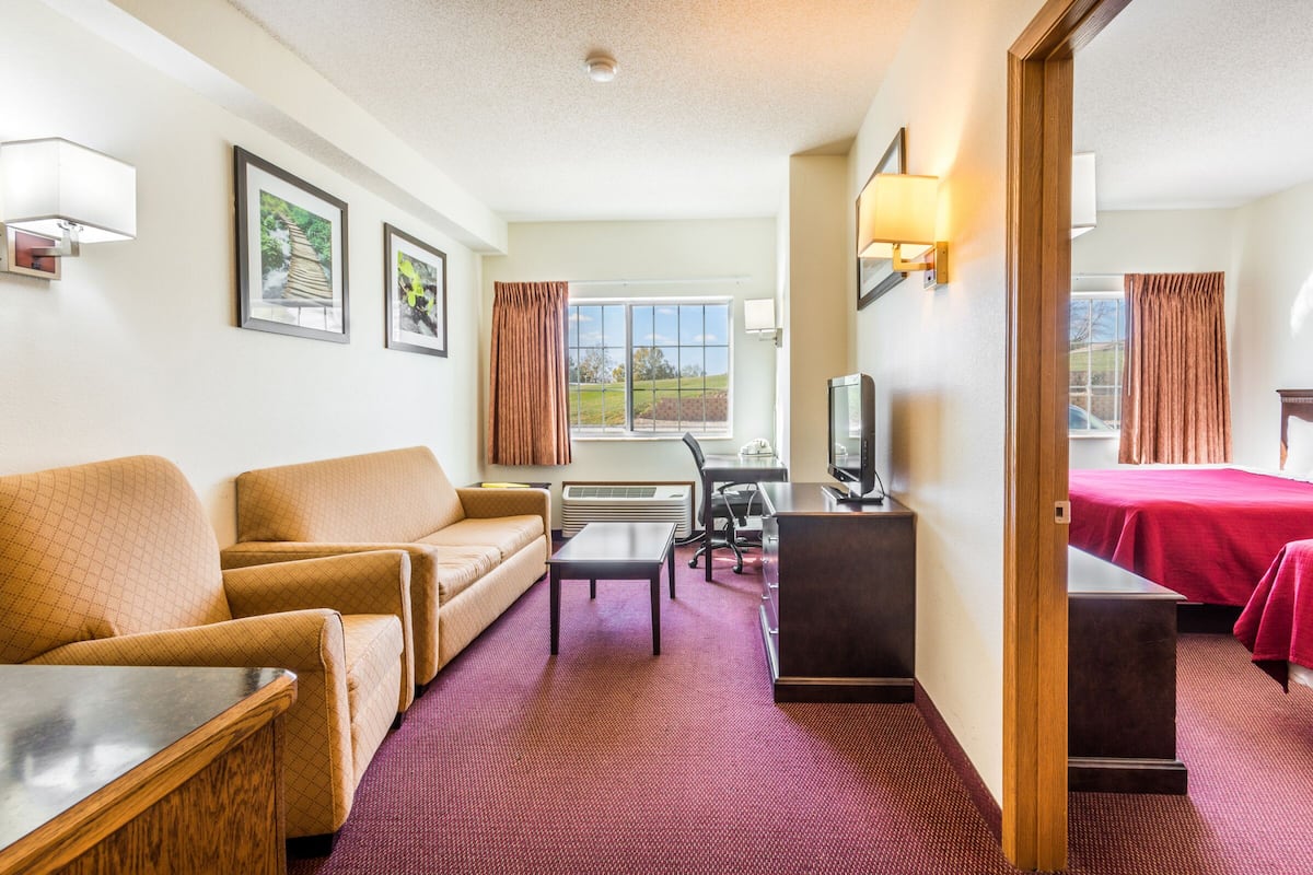 A hotel room with a sofa, a desk with a computer, a TV, and a bed with red bedding.