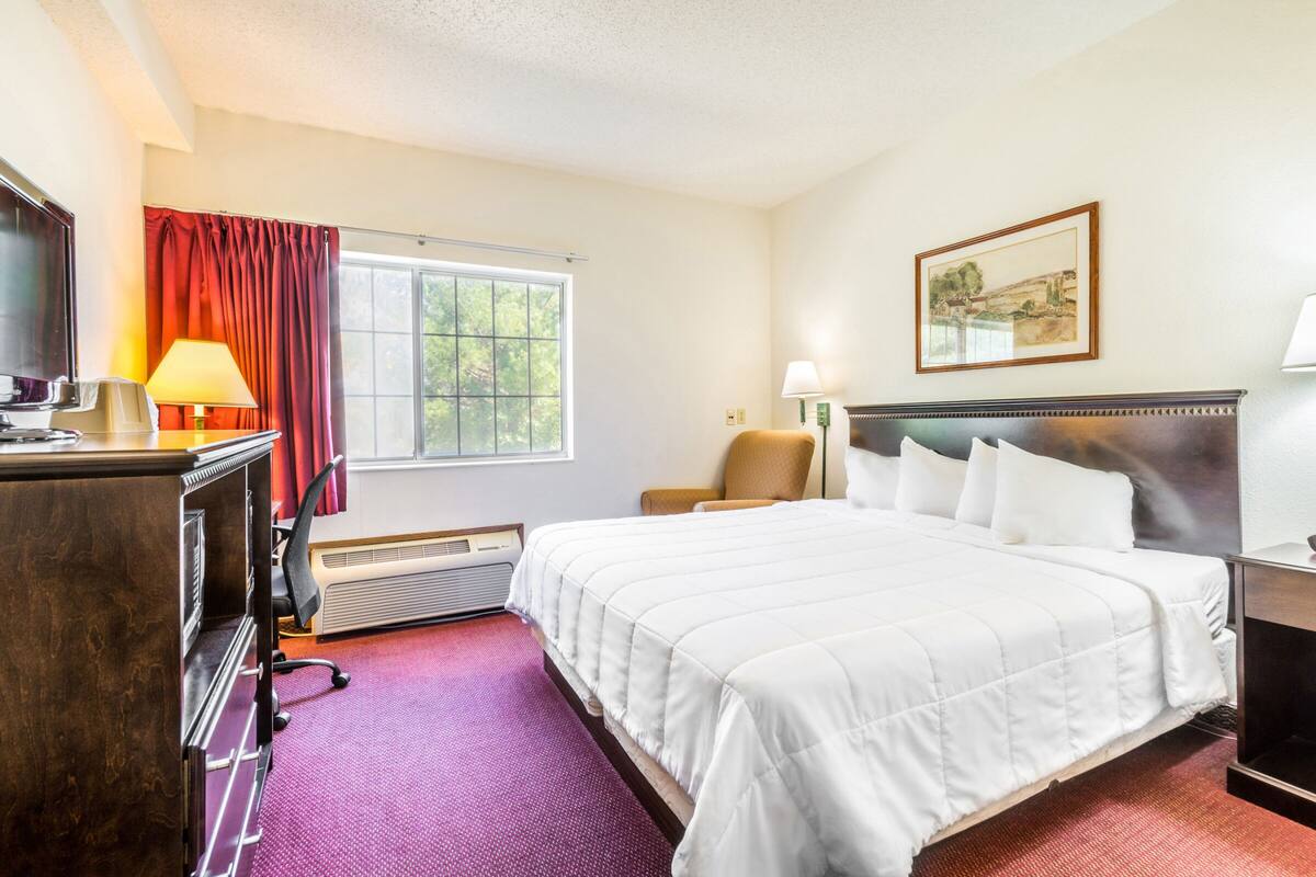 A hotel room with a large bed, a desk with a telephone, a chair, a window with curtains, a painting on the wall, and a clock on the nightstand.