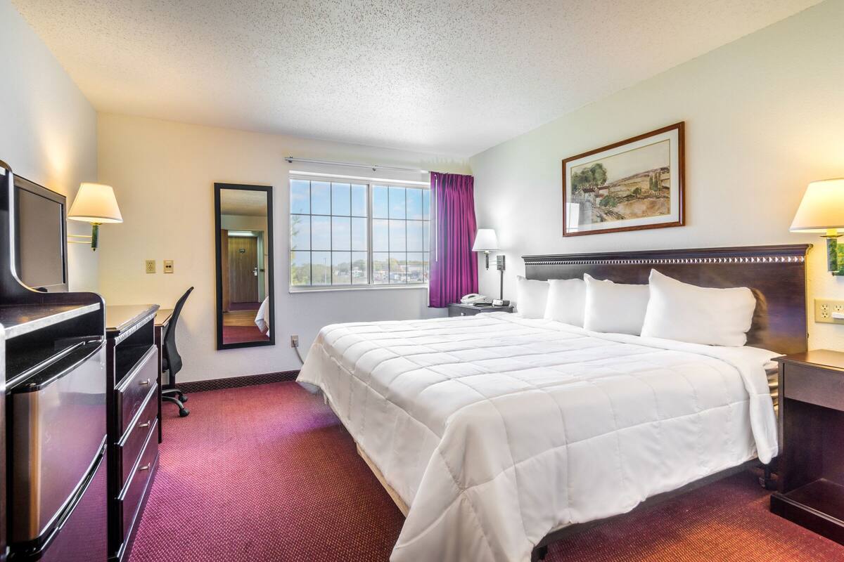 A hotel room with a bed, a desk with a computer, a TV, a mirror, and a window with curtains.