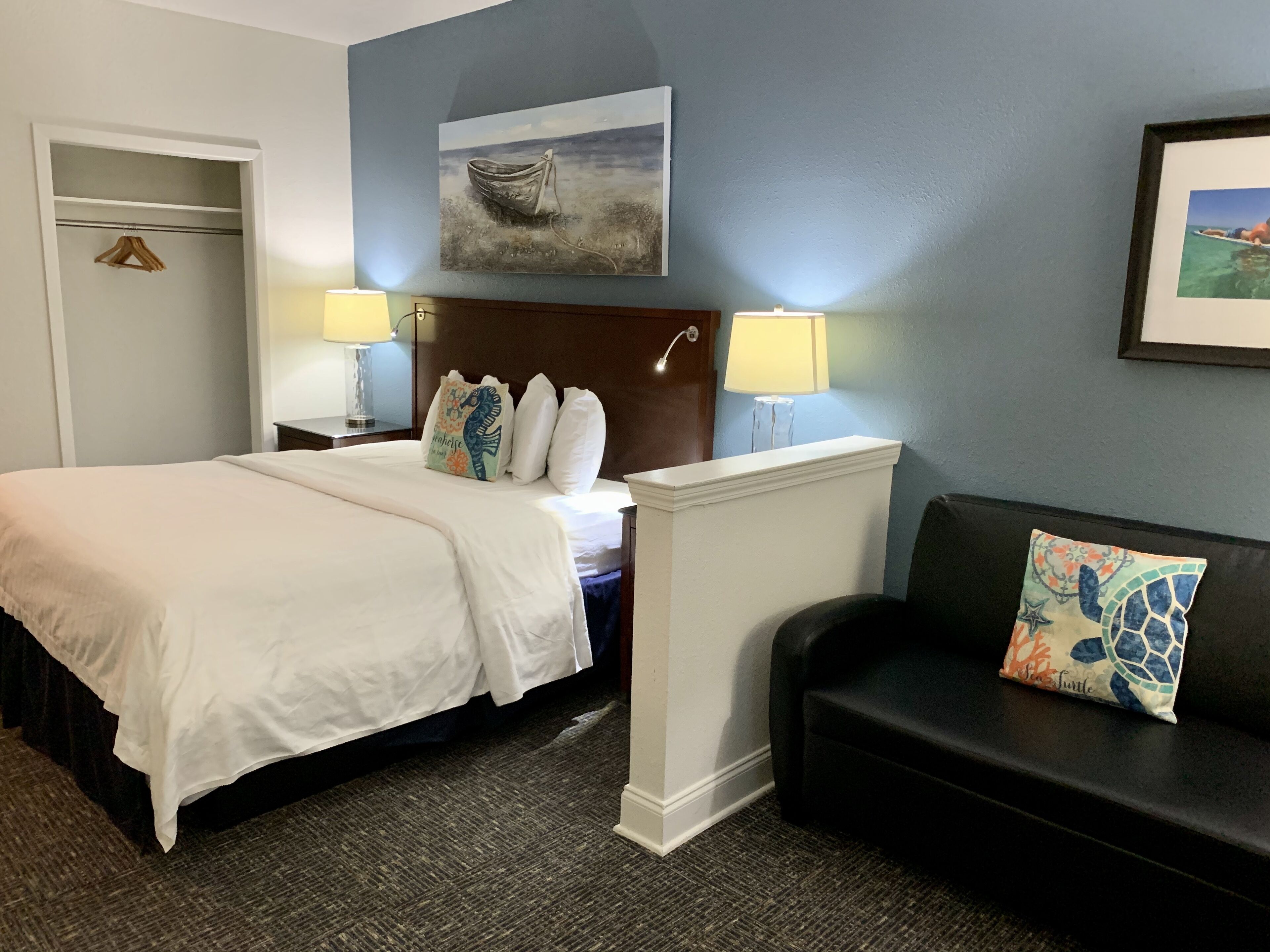 A hotel room with a bed, bedside lamps, a headboard, a painting, a framed picture, a black couch with a cushion, and a carpeted floor.