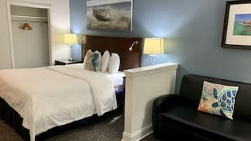 A hotel room with a bed, bedside lamps, a headboard, a painting, a framed picture, a black couch with a cushion, and a carpeted floor.