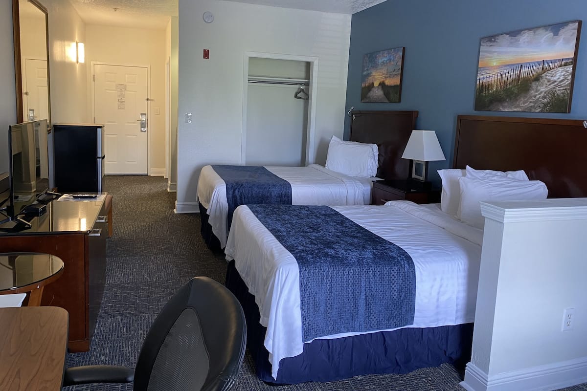 A hotel room with two beds, a desk with a computer, a chair, and a TV. There is a door and a coat rack in the background.