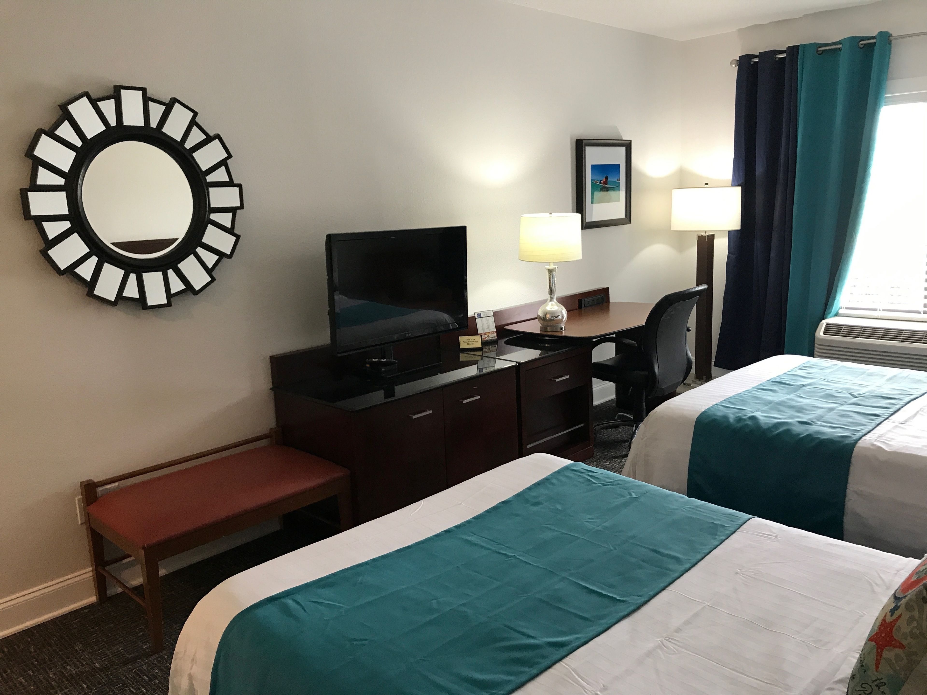 Habitación de hotel con dos camas, un escritorio con televisor, un espejo, una lámpara y una ventana con cortinas.