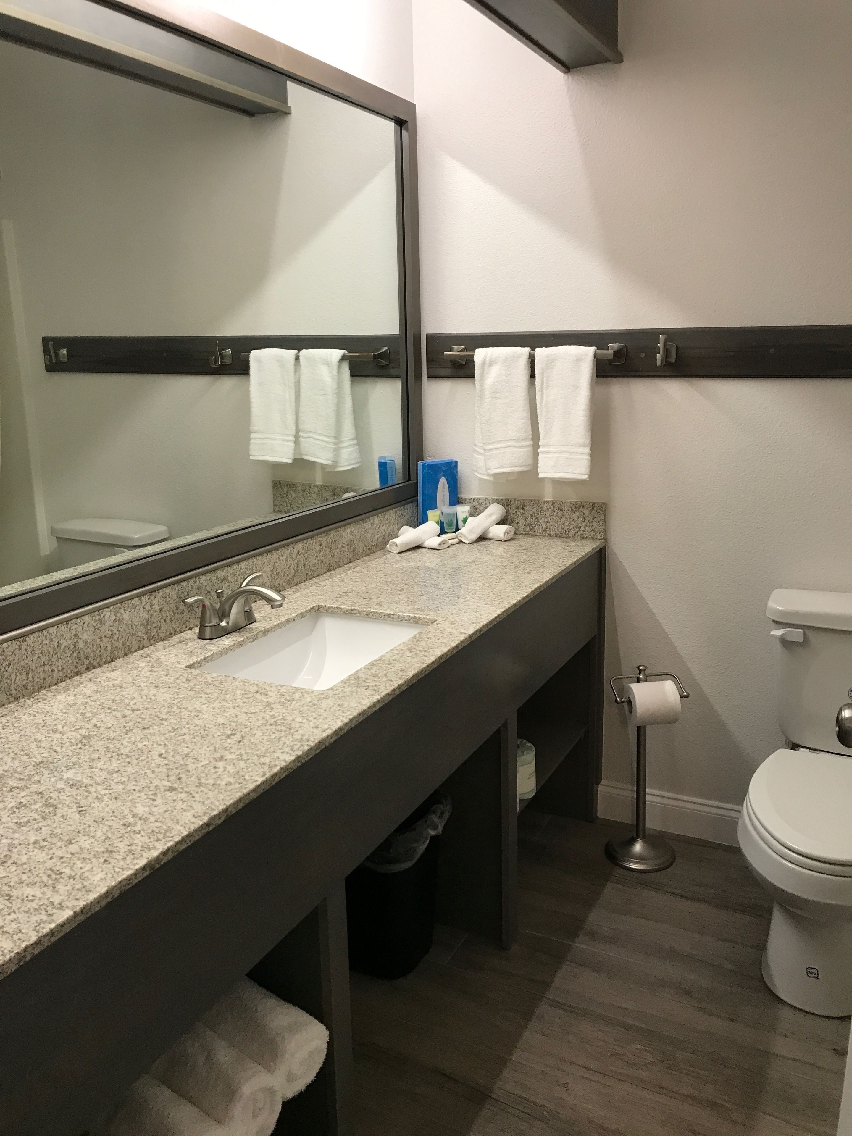 A modern bathroom with a granite countertop, two sinks, a large mirror, a toilet, and a towel rack.