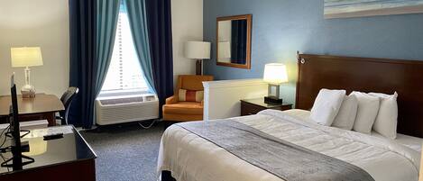 A hotel room with a bed, a desk with a computer, a chair, a lamp, a window with curtains, and a painting on the wall.