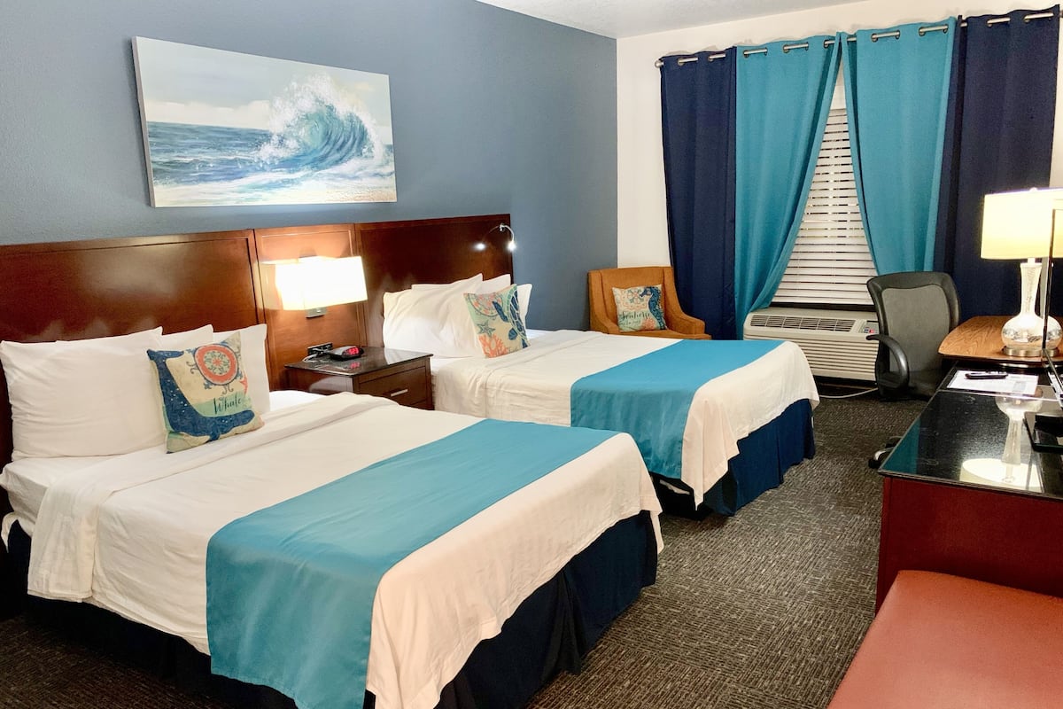 A hotel room with two beds, a desk, a chair, a desk lamp, a painting of a wave, and a window with curtains.