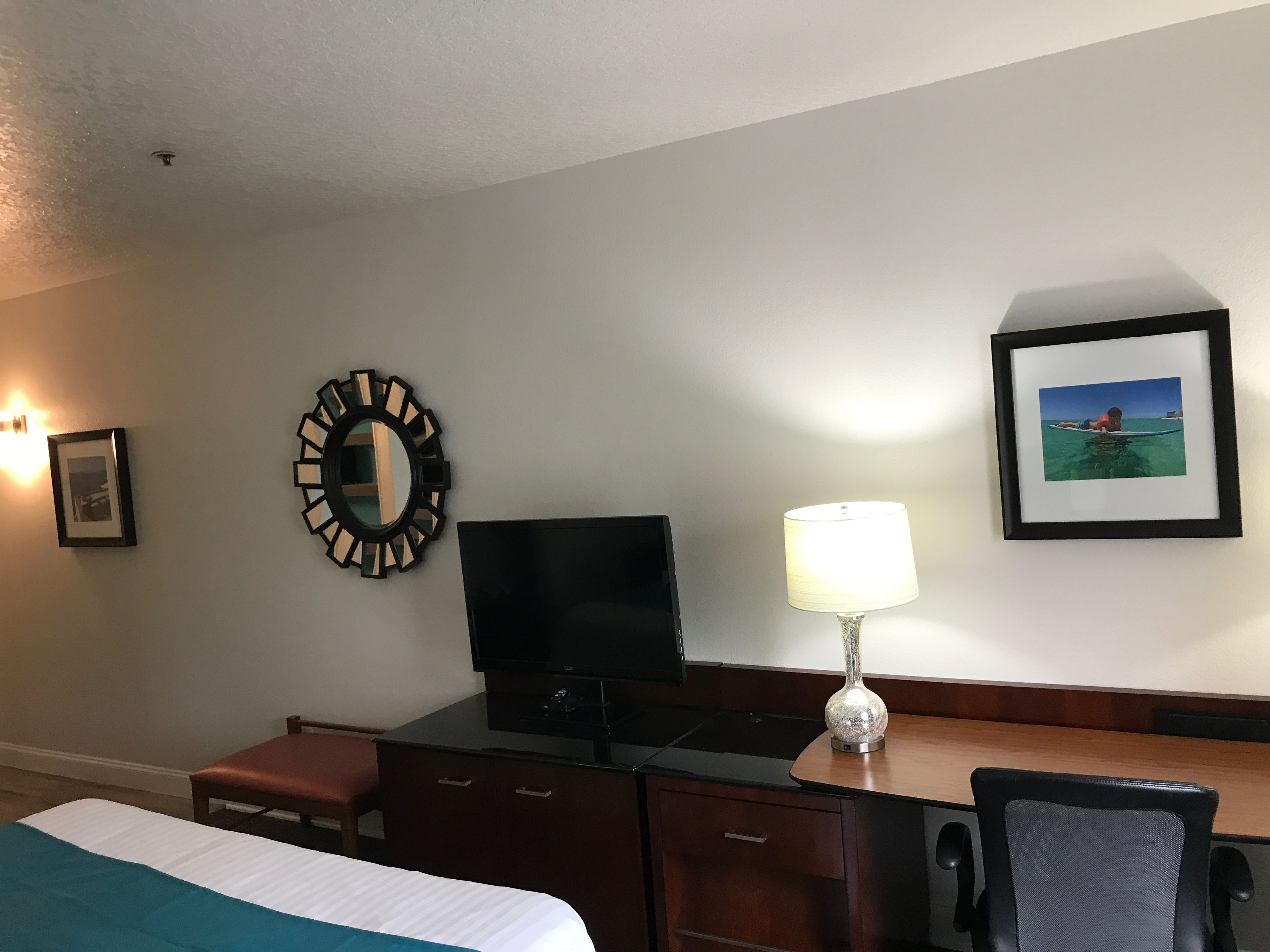 A hotel room with a bed, a desk, a television, a mirror, a lamp, and framed pictures on the wall.