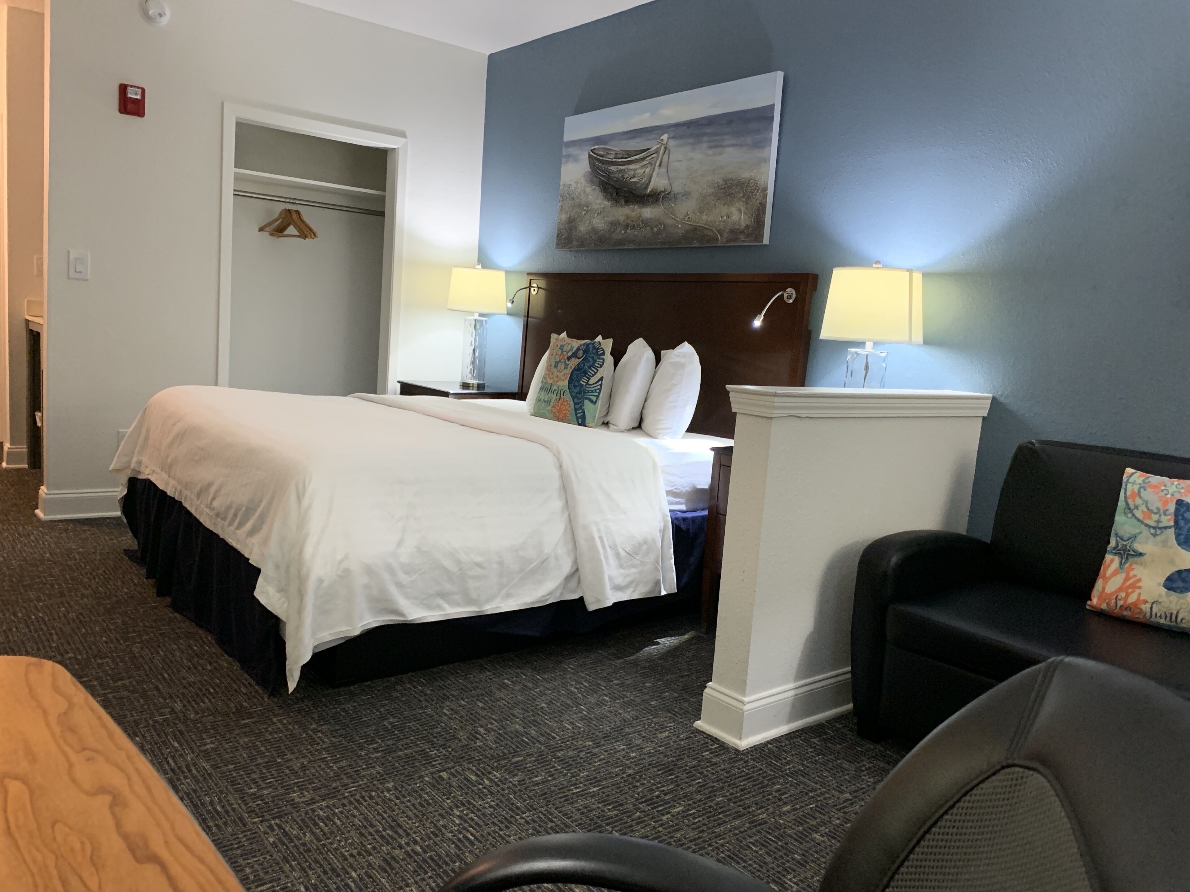 A hotel room with a bed, a desk, a chair, a coat rack, and a painting on the wall.