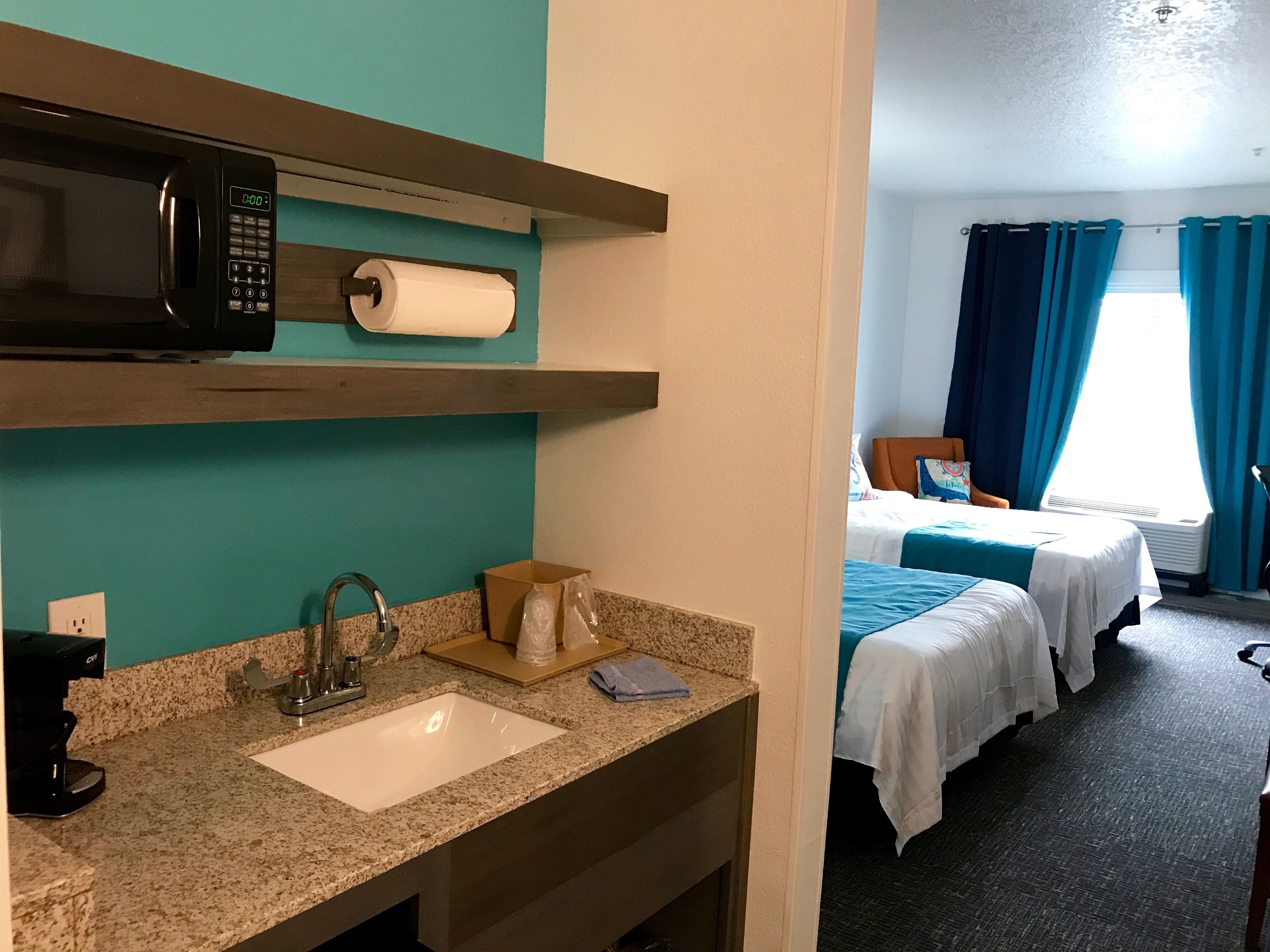 A hotel room with a kitchenette, a sink, a microwave, a toilet paper roll, a coffee maker, and a bed with blue bedding.