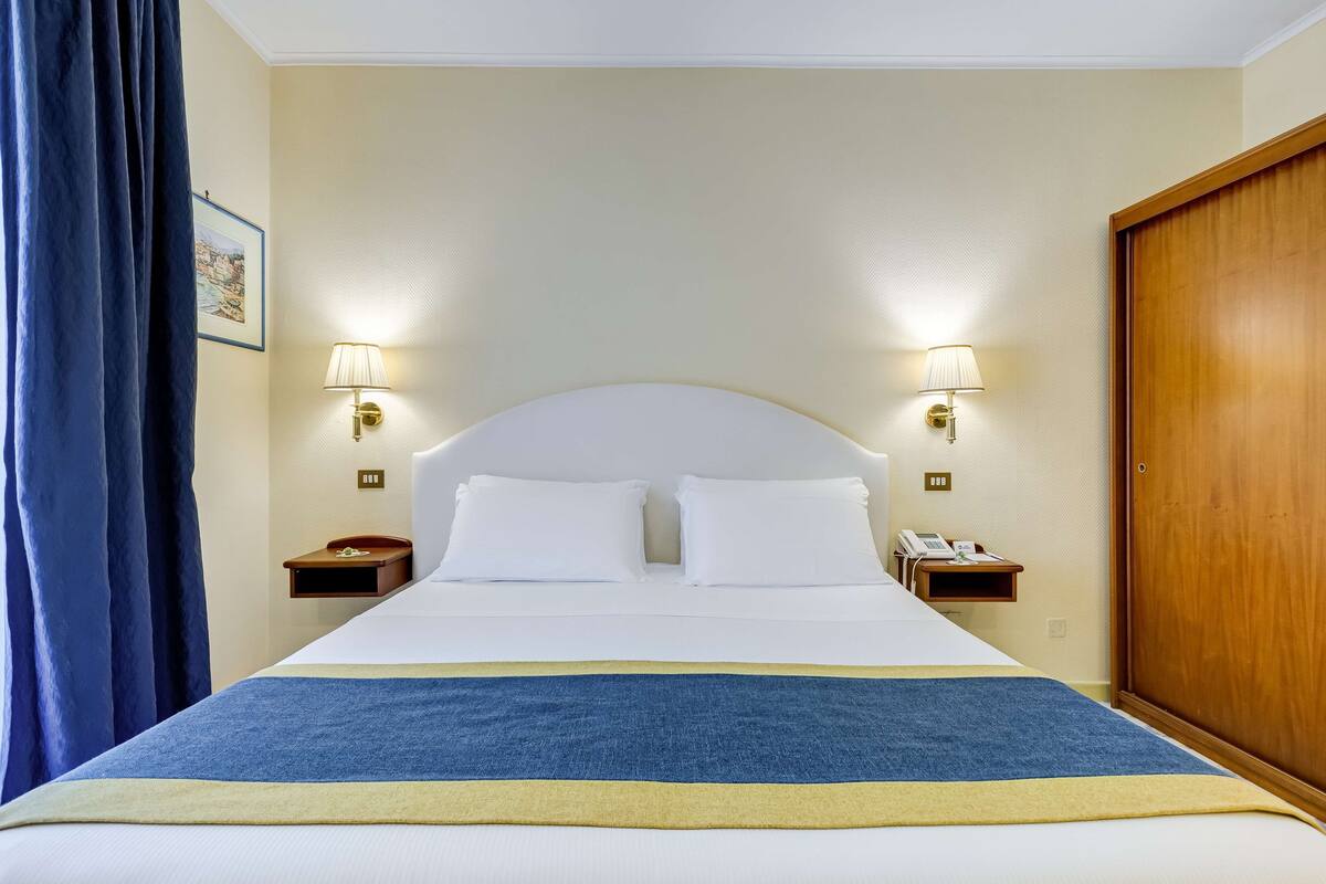 Een hotelkamer met een groot bed, twee bedlampen, een houten garderobe en een blauw gordijn.