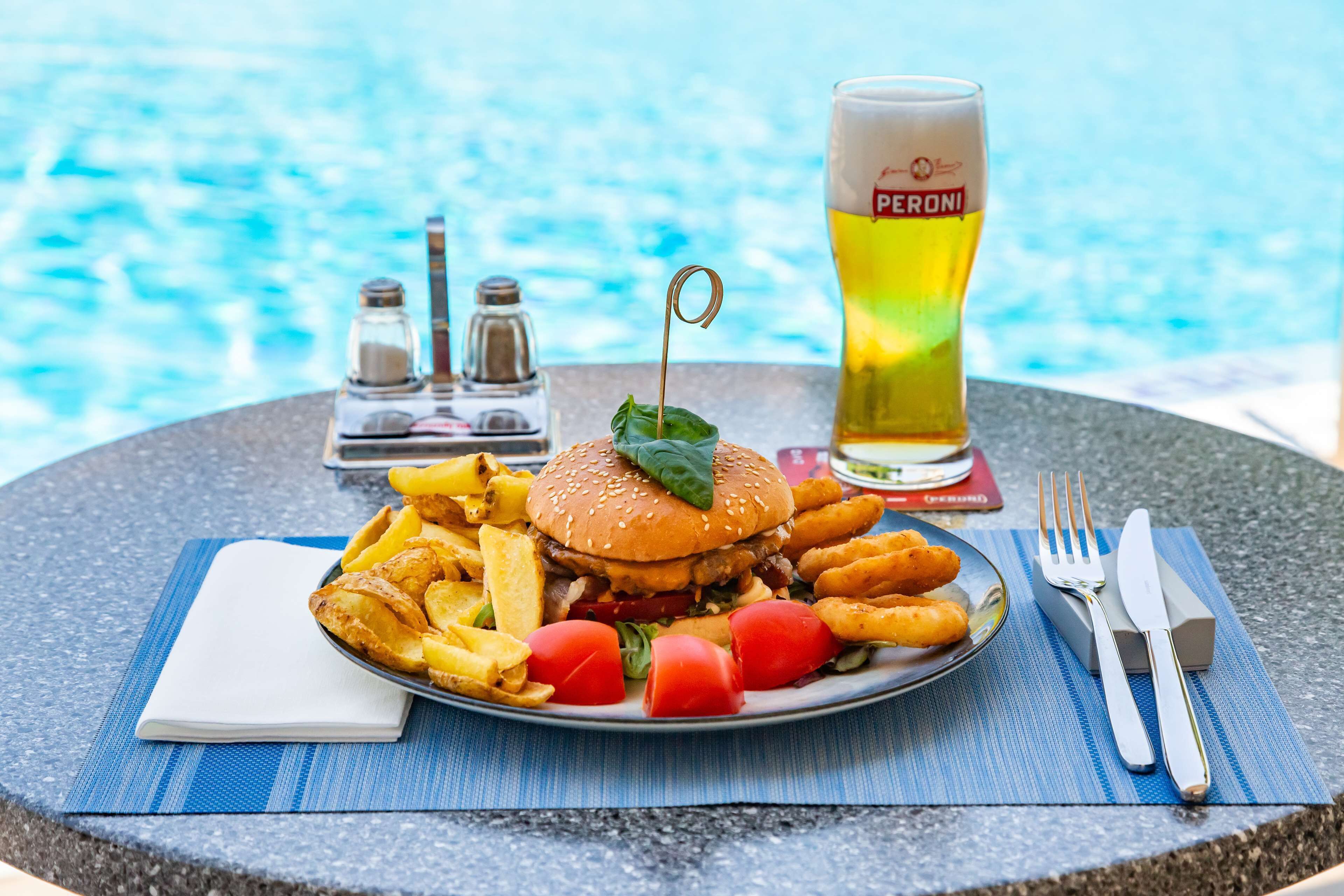 En måltid bestående av en hamburgare, pommes frites och ett glas öl, avnjuten vid ett bord intill poolen.