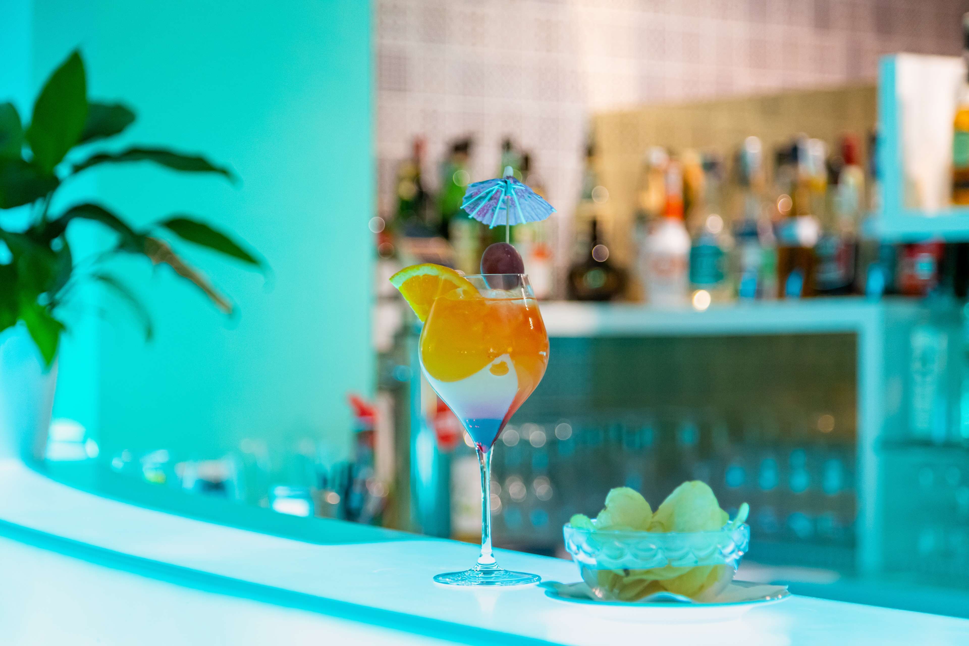 Ein Cocktail mit blauer Schirmchenverzierung und Zitronenscheibe, eine Obstschale und eine Bar mit Flaschen im Hintergrund.