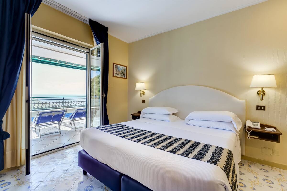 Hotelkamer met een groot bed, twee bedlampen, een nachtkastje met een telefoon, en uitzicht op het strand door een schuifdeur.