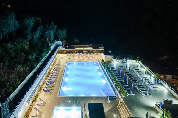 A large indoor swimming pool with multiple lanes, surrounded by lounge chairs and umbrellas, and a covered seating area with tables and chairs.