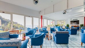 Een ruime zithoek met blauwe banken en stoelen, grote ramen met uitzicht op de omgeving, en een discobal aan het plafond.