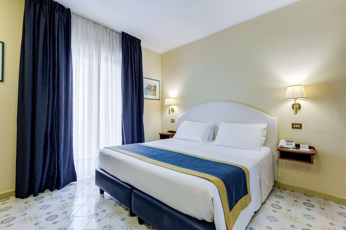 Een hotelkamer met een bed, gordijnen, een nachtkastje met een lamp en een aan de muur bevestigd telefoon.