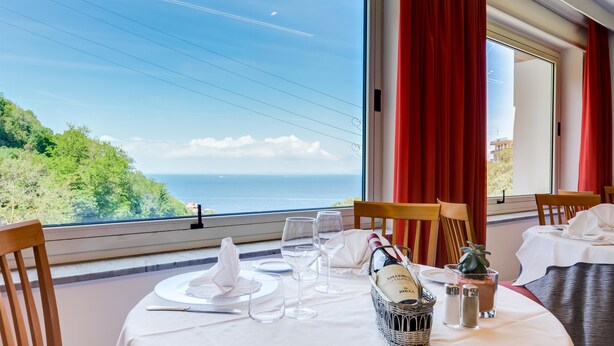 Een eettafel met uitzicht op zee en heuvels, rode gordijnen en een fles champagne.