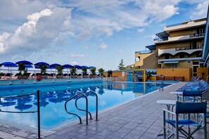 Une piscine avec des chaises longues et des parasols, une terrasse carrelée et un bâtiment doté de balcons.