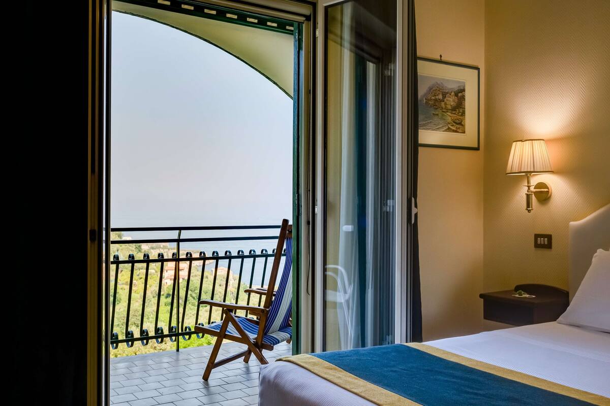 Een hotelkamer met een bed, nachtkastje, lamp, stoel en een balkon met uitzicht op zee.