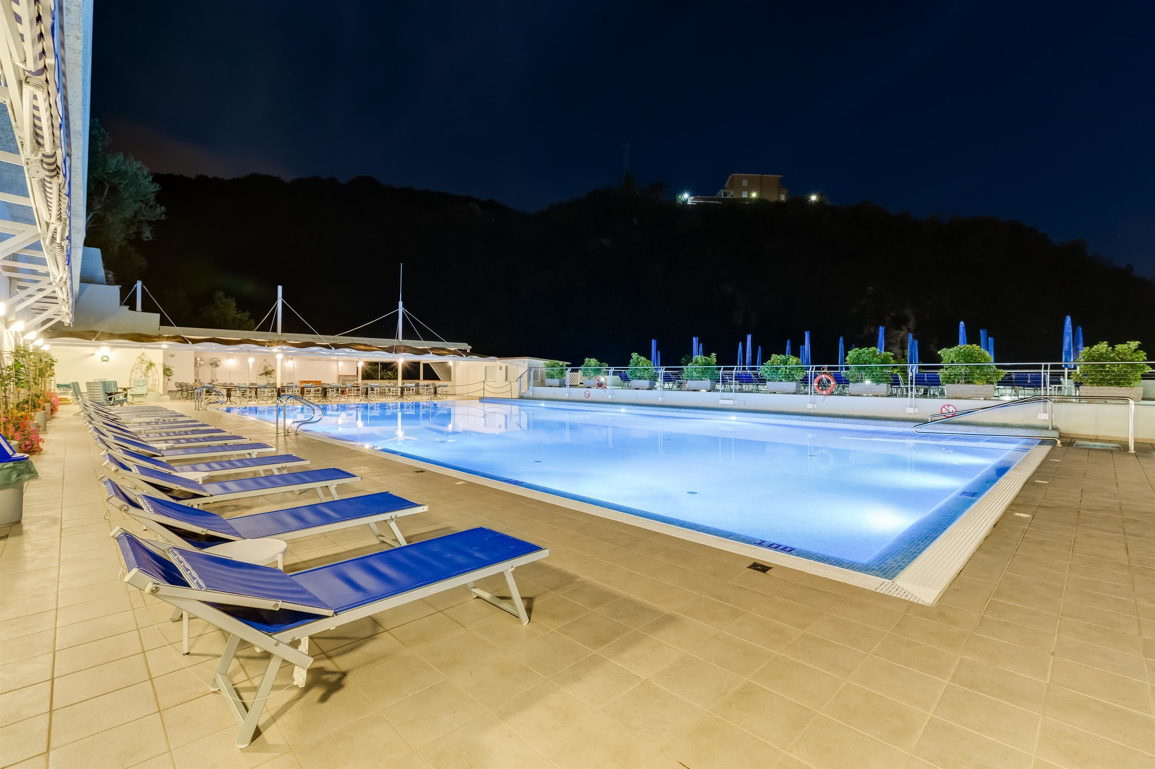 Piscina con sillas de descanso y sombrillas, rodeada por un edificio con vistas a una colina.