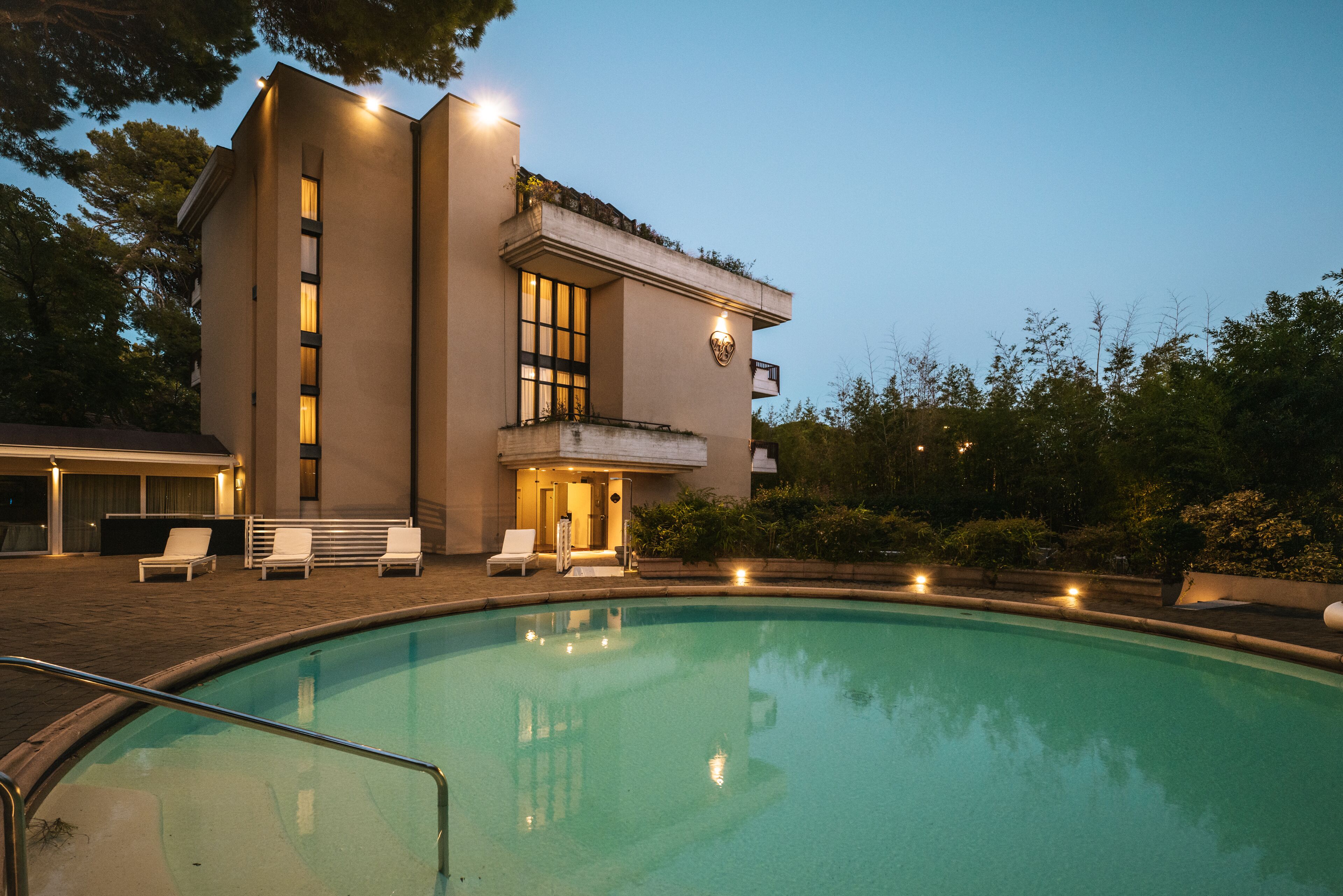 A modern building with a swimming pool, outdoor seating, and a well-lit entrance.