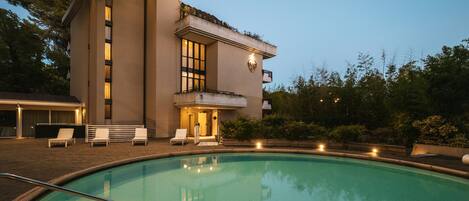 A modern building with a swimming pool, outdoor seating, and a well-lit entrance.