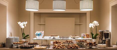 A buffet setup with various baked goods and pastries, including cakes and croissants, under three striped pendant lights.