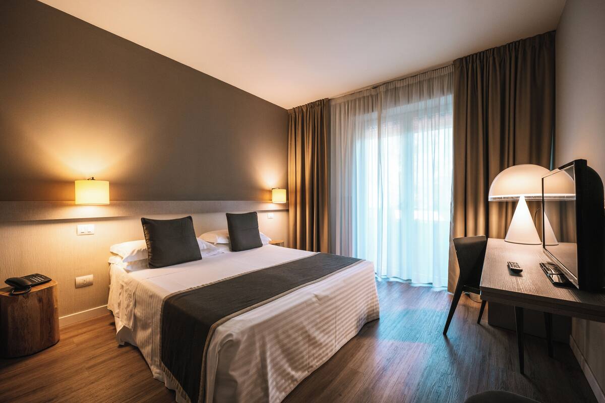 Een hotelkamer met een bed, nachtlampjes, een bureau met een lamp en een stoel.