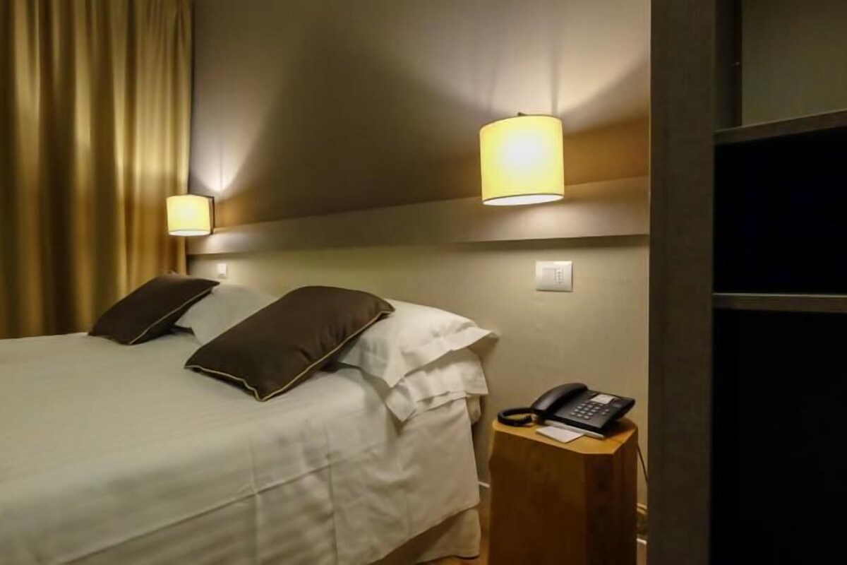 A hotel room with a bed, two pillows, a bedside table with a phone, and wall-mounted lamps.