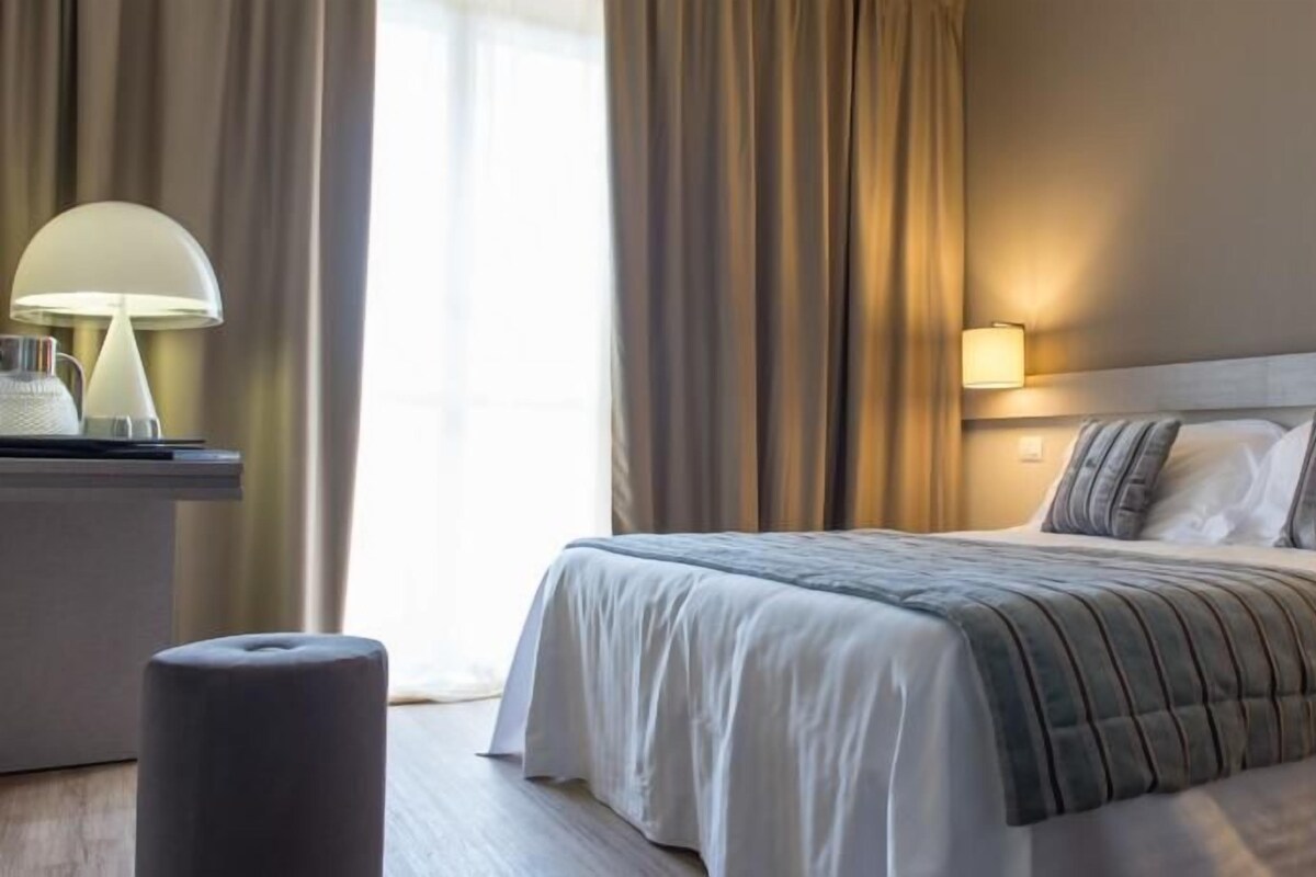 Une chambre d’hôtel avec un lit bien fait, une lampe de chevet, une petite table avec une lampe et une fenêtre avec des rideaux.