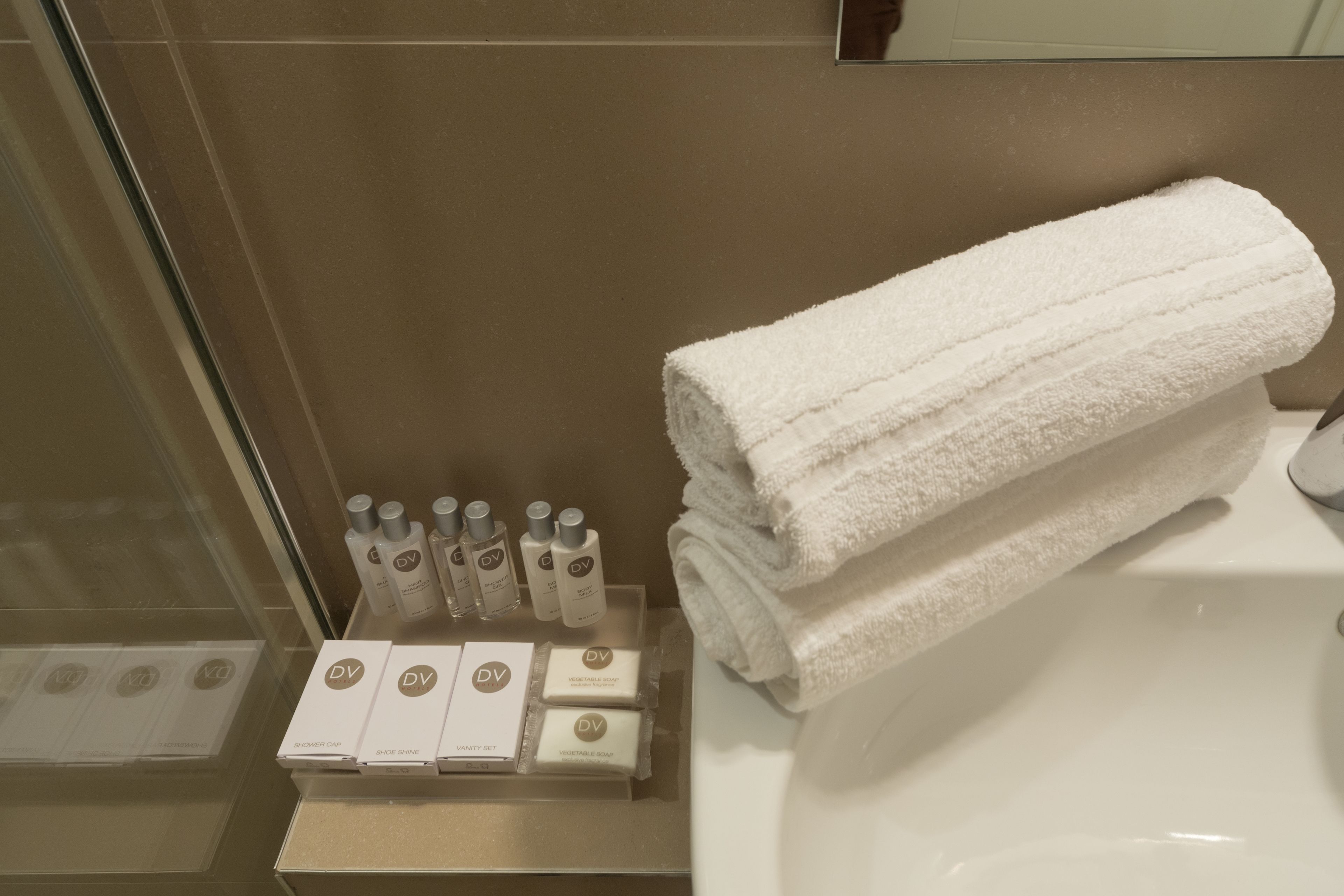 Un baño con una toalla blanca doblada sobre un lavamanos blanco, y un estante con varios artículos de aseo, incluyendo un gorro de ducha, kit de limpieza de calzado y un set de tocador.