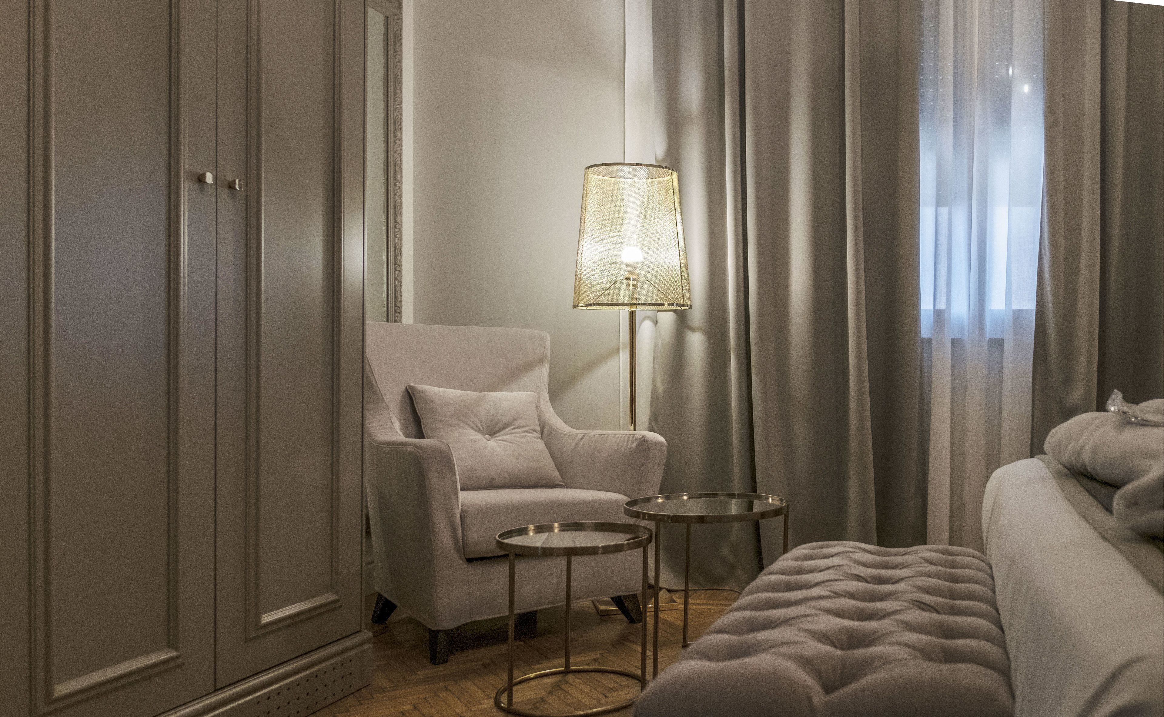 A hotel room with a large bed, a sitting area with a chair and a small table, a floor lamp, and a wardrobe.