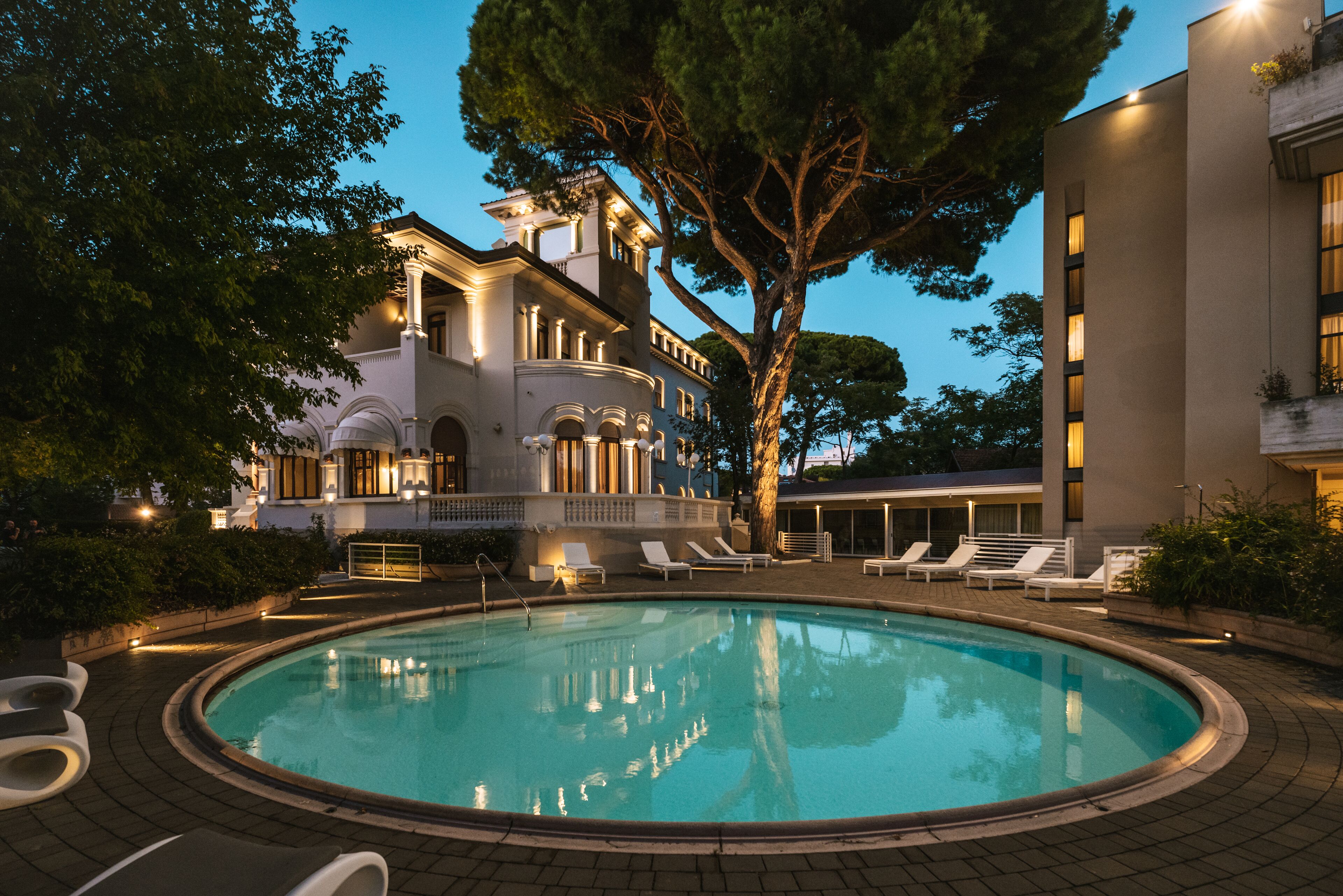 A swimming pool with lounge chairs, a large tree, and a building with classical architecture.