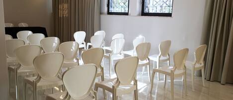A conference room with white chairs arranged in rows, large windows with black frames, and beige curtains.