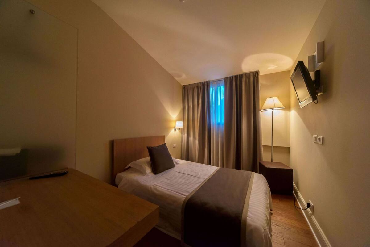 Een hotelkamer met een bed, nachtkastje, lamp en een aan de muur bevestigde televisie.