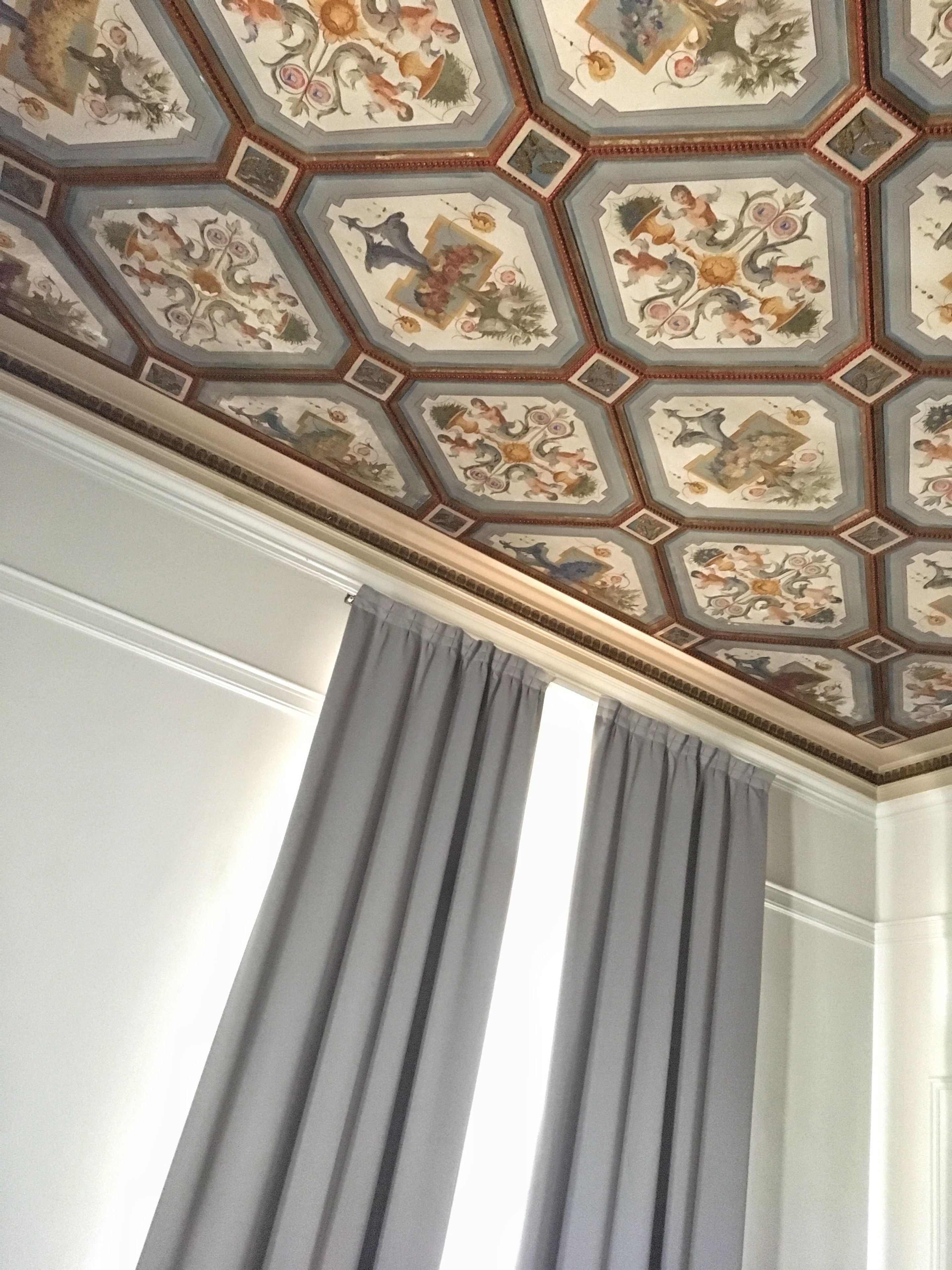 A room with a ceiling featuring a geometric pattern with floral and animal motifs. There are two large windows with grey curtains.