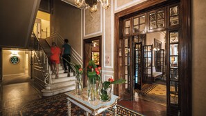 A grand lobby with a marble staircase, a chandelier, and a large wooden door leading to a room with a bed.