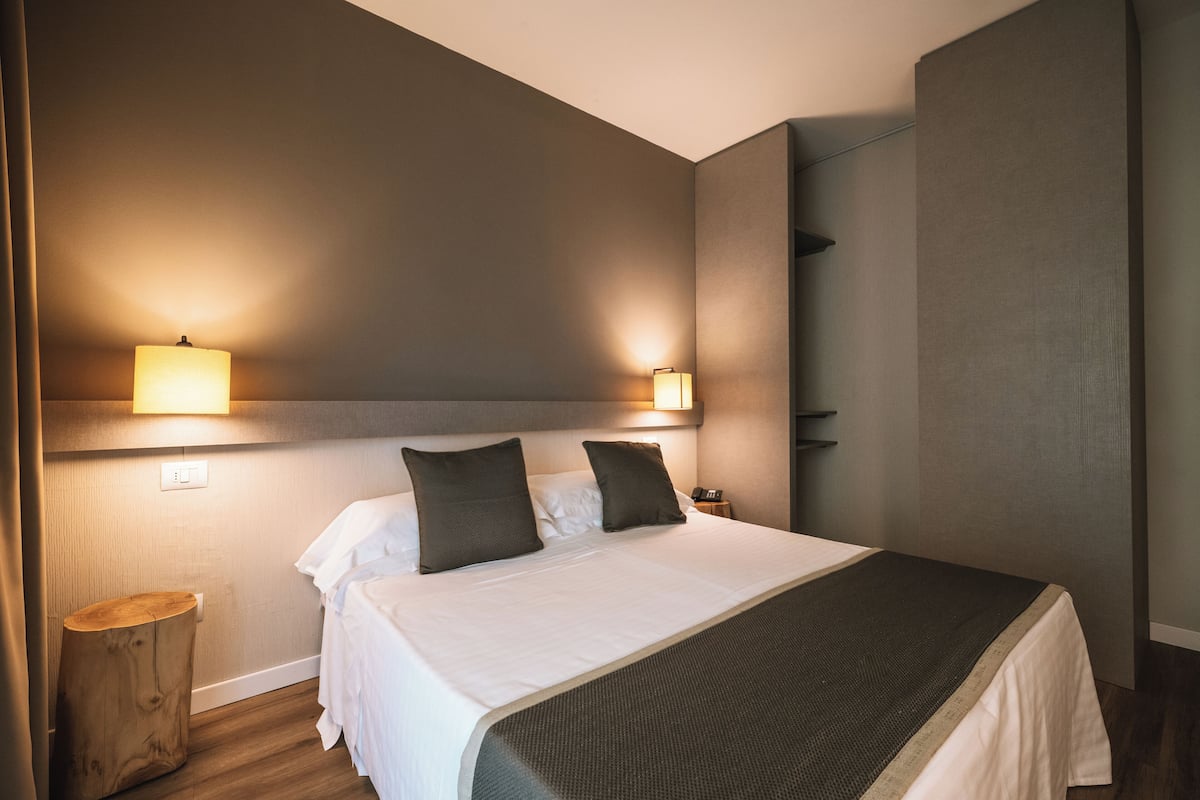 Een opgemaakt bed met een witte dekbedovertrek en donkere kussens, een nachtkastje met een lamp, en een klein houten krukje.