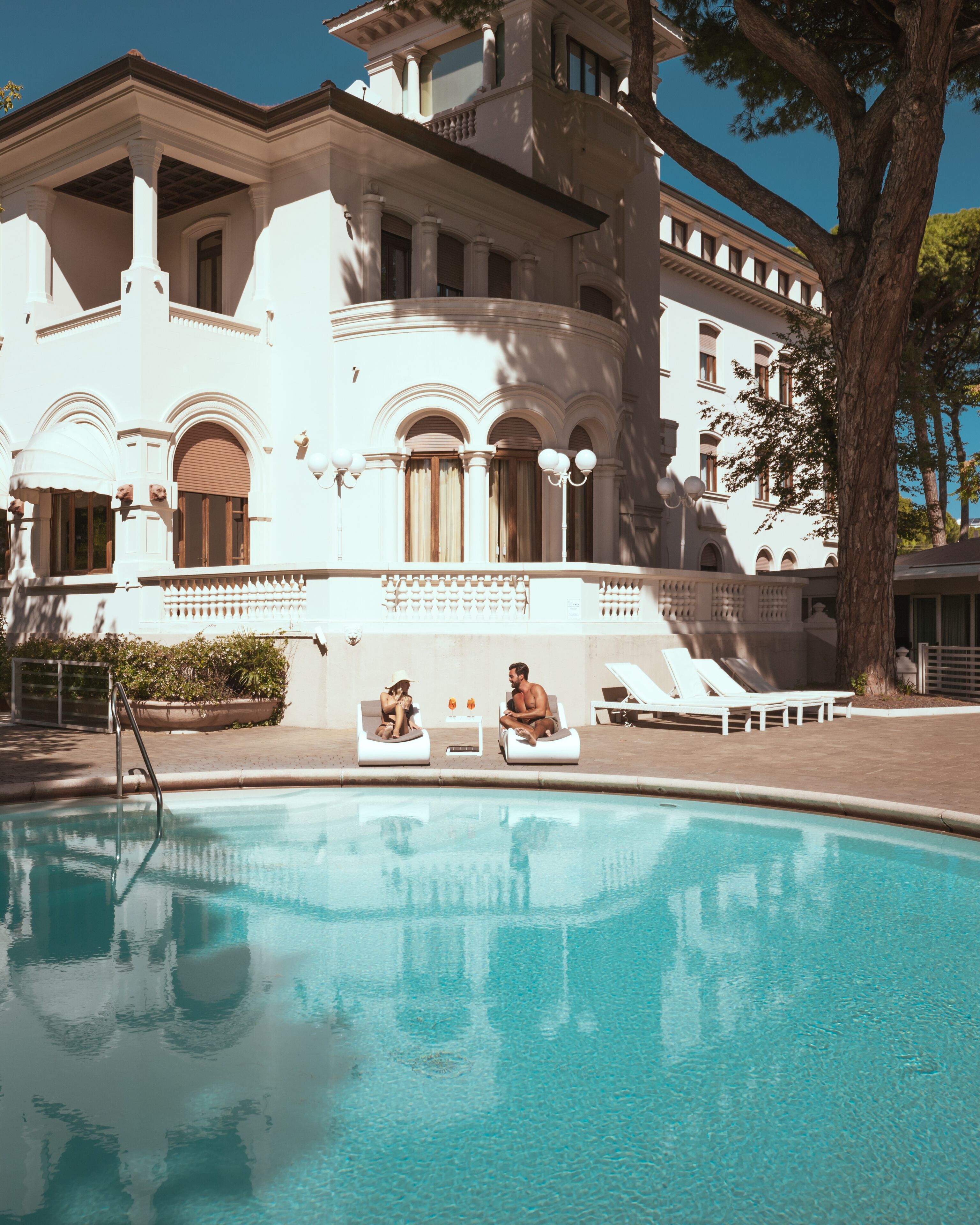 Zona de piscina con sillones de descanso y un edificio de arquitectura clásica.