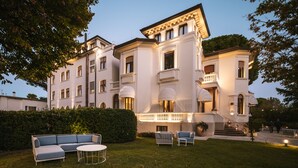 A large, white, multi-story building with a formal architectural style, including a balcony and a well-lit entrance.