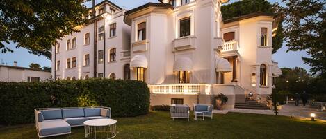 A large, white, multi-story building with a formal architectural style, including a balcony and a well-lit entrance.