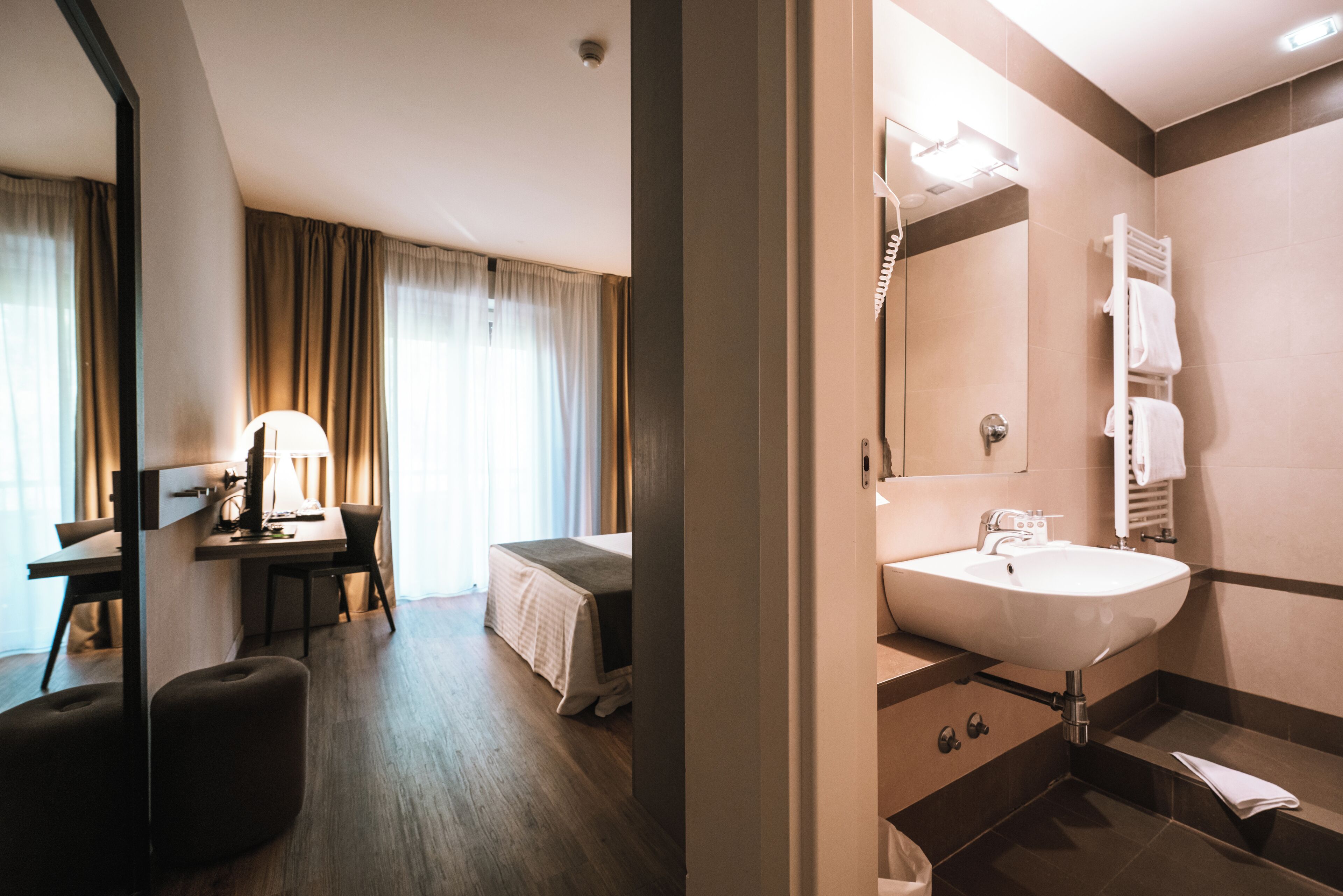 A hotel room with a desk, chair, bed, and a bathroom with a sink and mirror.