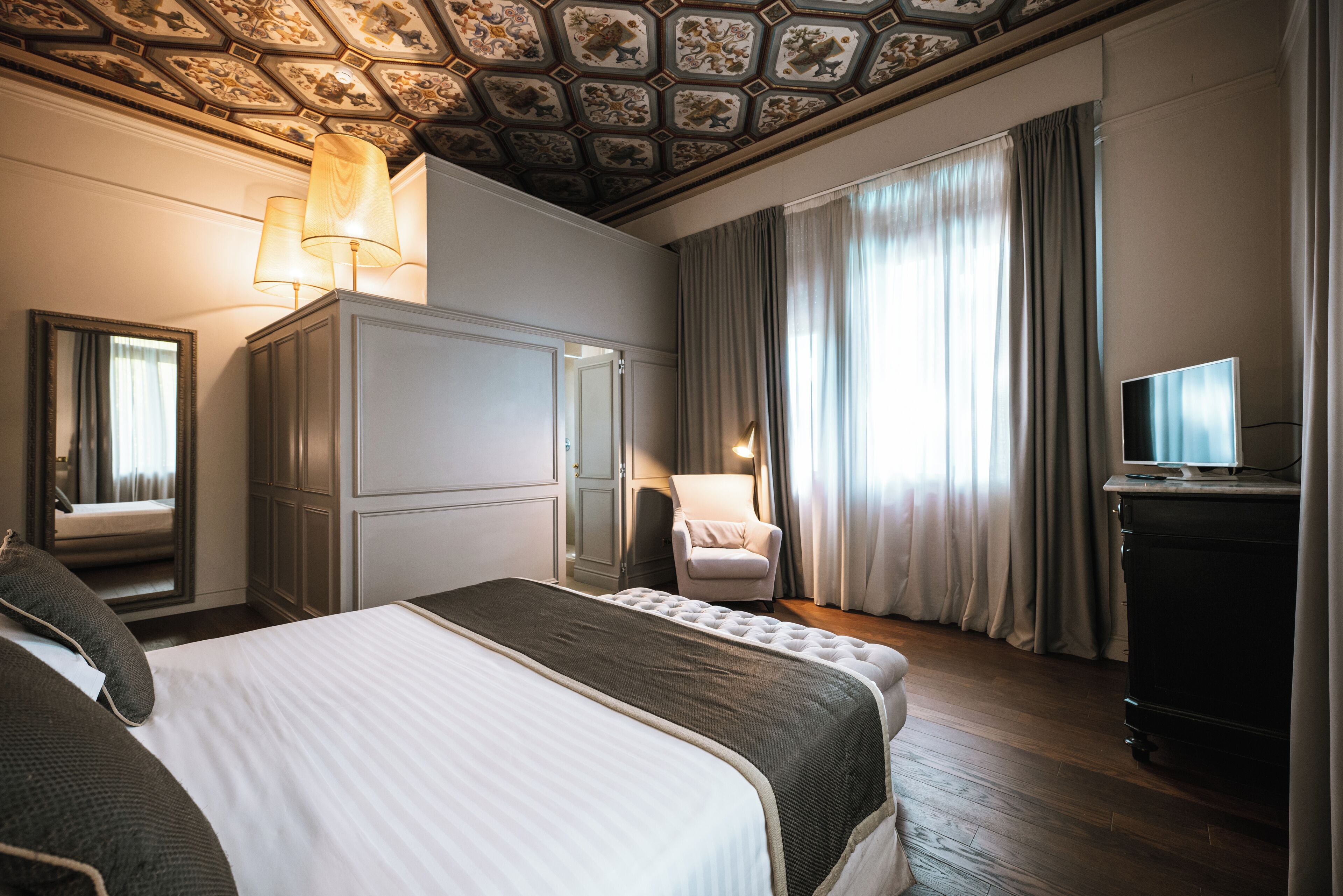 Habitación de hotel con una cama grande, un armario, una silla, un televisor y un techo con diseño decorativo.