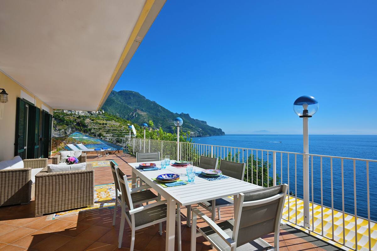 Une terrasse avec une table dressée pour le dîner, offrant une vue sur la côte et les montagnes en arrière-plan.