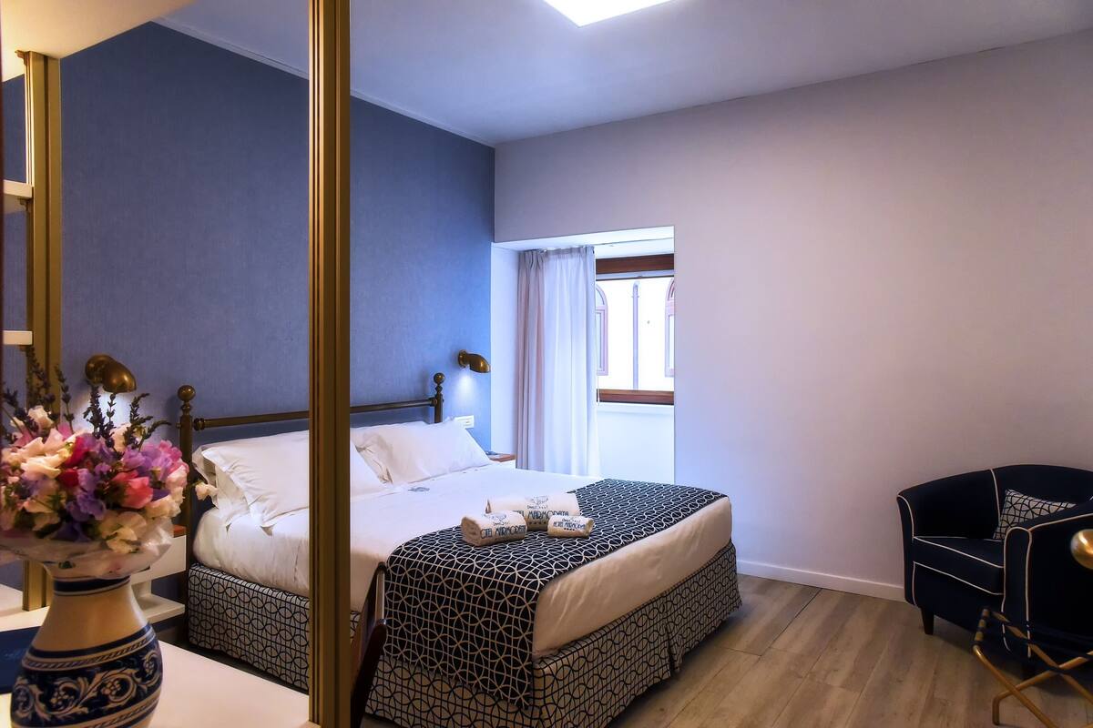 Une chambre d’hôtel avec un lit, une table de chevet, un vase de fleurs, une chaise et une fenêtre avec des rideaux.