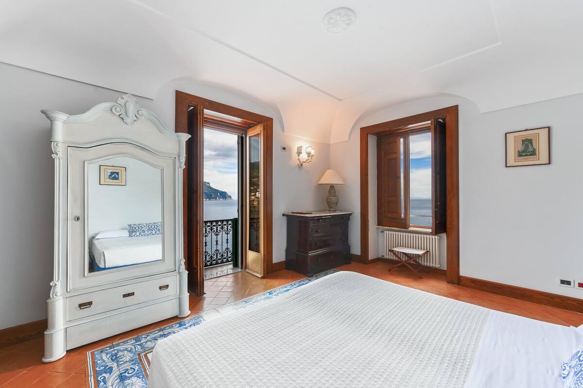 Une chambre à coucher avec un grand lit, une commode, une lampe et une vue sur la mer par la fenêtre.
